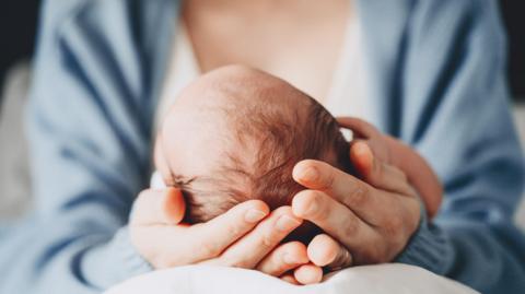 A mother resting a baby's head in the palm of her hands, on her lap, so that we can see only the back of the infant's head. The mother is wearing a blue cardigan.