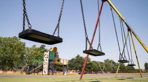 A row of swings in an outdoor grassy area. Behind them, further away, is a large climbing frame with slide, climbing wall and green and yellow frame. Behind that, a row of trees.
