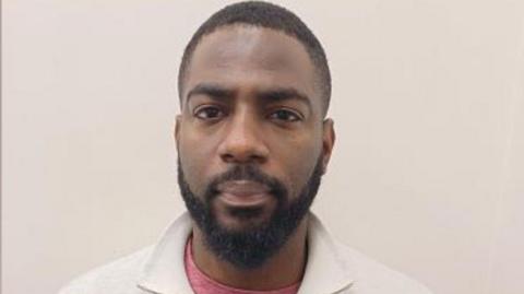 A man's mugshot. He wears a white jumper with a pink top underneath. 