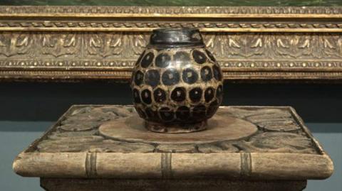 The stoneware jar on a plinth. The jar has a tortoiseshell design. It is round and dark brown in colour.