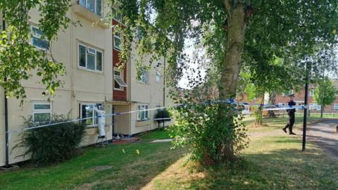 A sunny grass verge outside a block of flats cordoned off by blue and white police tape with a forensic officer and a police officer at the scene.