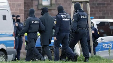 Seen from behind, a group of police officers with POLIZEI written on their backs escort a man, covered in dark clothing and something covering his face, towards a police van.