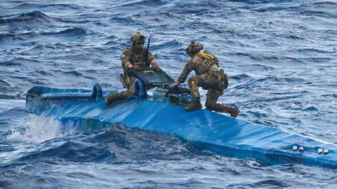 Two personnel in camouflage outfits squat on top of a semi-submerged long, blue vessel in the sea