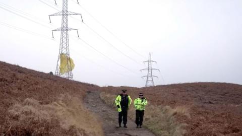 Parachute stuck in pylon on moors - with two police officers walking down hill dressed in yellow jackets.