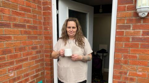Hollie, a woman in her 40s with long hair, stands outside her back door with a cup of tea.