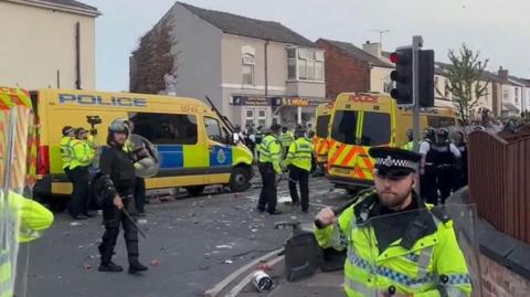 Police officers some in riot gear dealing with disorder in Southport on 30 July. Uniformed officers can be seen holding riot shields. There are two yellow police vans and a man in dark clothing holding a shield and a baton. Debris is strewn across the road. 