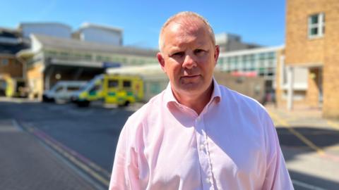 Steve McManus has short grey hair and is wearing a pink shirt. He's standing outside on a sunny day, with a hospital and an ambulance in soft focus behind him.