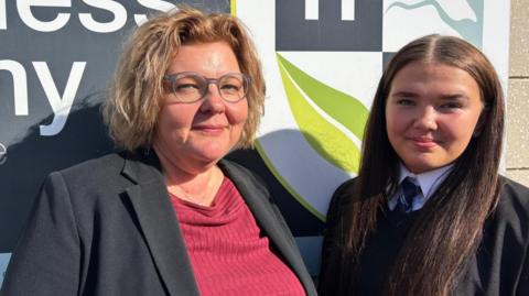 A woman with short blonde hair and lilac glasses is wearing a black blazer and pink top and smiling into the camera. Next to her is a teenage girl wearing a school uniform and tie with long brown hair smiling into the camera. 