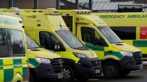 Five ambulances are lined up outside a hospital. The red 999 logo can be seen on the side of the vehicle