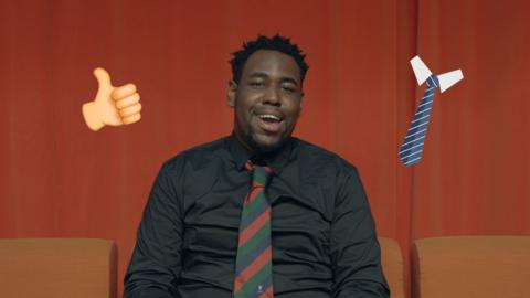 BBC journalist Terel Edmunds sat in front of an orange background. He is smiling at the camera and wearing a black shirt with a stripey tie.