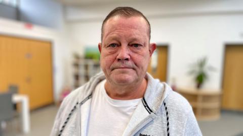 Gary James is wearing a grey hooded sweatshirt with a white T-shirt underneath. Behind him is a bookcase and a plant in the lounge of Elm House