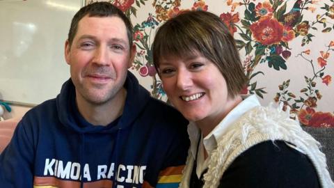 Stef Scurr and her husband Andrew Dunn smile at the camera, against a floral wallpaper background. Stef has short brown hair and wears a white gilet, white shirt and black jumper. Andrew has short brown hair and wears a navy hoodie with white writing across the chest.