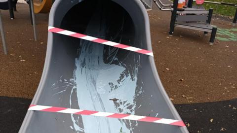 The bottom of a grey tube slide with blue paint splattered across it. There is red and white tape across the bottom of it. In the background you can see brown and black tarmac as well as other play equipment