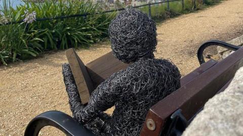 A metal sculpture of a human figure reading a book on a wooden park bench