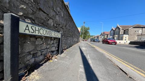 A residential road with a sign saying Ashcombe Road on it, and cars in the background