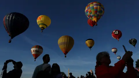 A group of colourful hot air balloons take off as people stand on the ground taking pictures of them