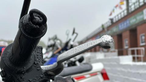 The black handle and silver brake lever of a motorcycle with droplets of rain gathered on them. A line of motorcycles parked in front of the grandstand can be see in the background.