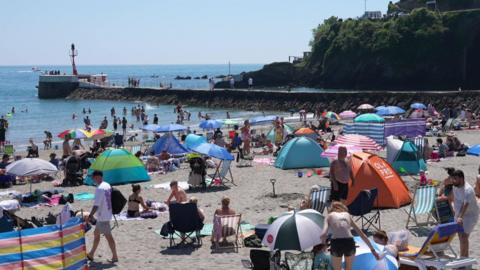 Lots of people enjoying Looe beach in Cornwall. There are deck chairs, people sat on towels and people swimming in the sea. The sea is in the background with clear blue skies. 