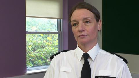 A police officer wearing uniform. She is looking directly at the camera. Her hair is tied back. Behind her is a window. There are bushed outside the window. 