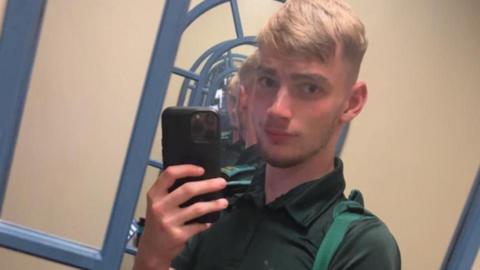 Jamie Kadolski stands in front of a mirror taking a selfie. He has a black phone up beside his face, and is looking directly into the mirror. He is wearing a green shirt. 