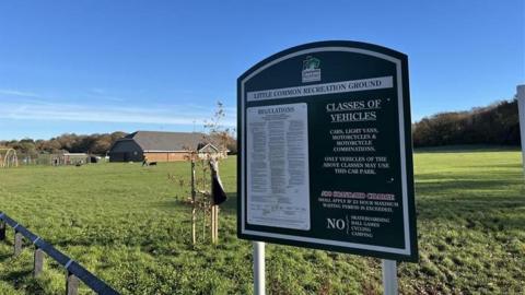 The sign for the recreation ground car park in the foreground with terms and conditions on it. In the background is the park and sports centre which cricket and football teams use