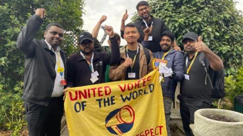 Seven members of the cleaning and porters team at St Helier and Epsom Hospital hold up a yellow banner with the name of their union, United Voices of the World, printed on it, as it each member of the group raises their hands in salute.