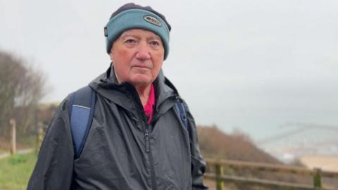 Derrick Downs wearing a blue woolly hat and waterproof jacket looking to camera with the coastline of Dover behind him