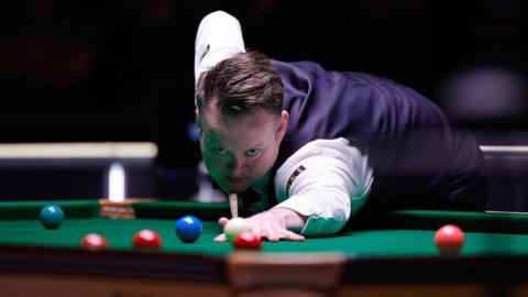 Shaun Murphy strikes the white ball towards a red. He has dark hair, brushed to his left, and is wearing a white shirt, a dark waistcoat and dark trousers. The baize on the table is green, and several balls can be seen - four reds, the green and the blue.