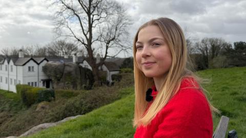 A young woman with long blonde hair in a bright red jumper sitting a bench overlooking a white house