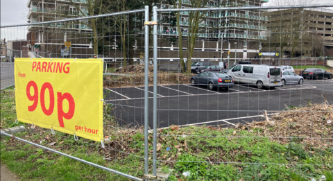 A yellow sign advertising 90p parking on a metal gate with several cars parked in spaces. 