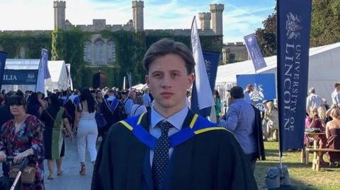 James Harrison at his graduation ceremony stood outside. He's wearing a black cape with blue and yellow accents. He is in front of a bustling crowd of other graduates.