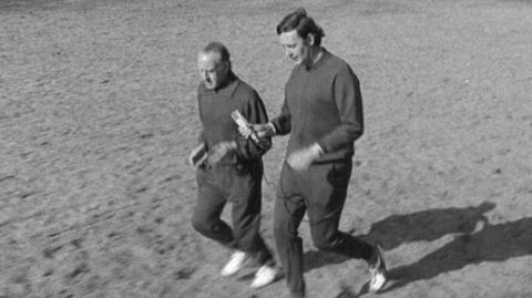 Two men wearing tracksuits jog across the grass in the park.