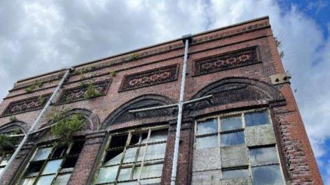 A red brick building with numerous broken window panes and vegetation growing up parts of the building.