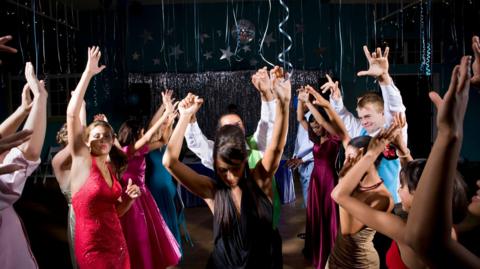 A mixture of male and female teenagers dancing in formal wear on a dance floor with a black backdrop with silver tassels and streamers. They all have their hands in the air and line up next to a couple in the middle.