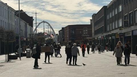 People walk along a pedestrainised street in Bolton town centre, lined with shops. A modern white arch features in the background.