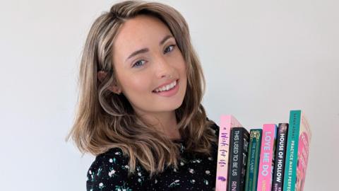 A woman holding books while smiling at the camera. She has a black top with a floral pattern on it.