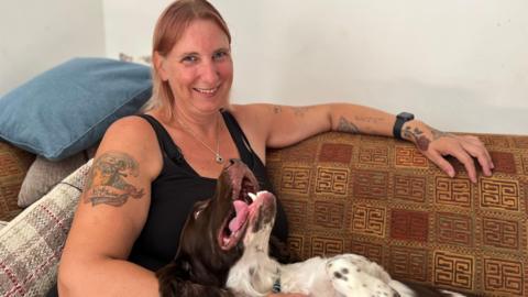 A woman with tattoos sitting on a brown sofa with her springer spaniel. She is looking ahead and smiling and her spaniel is looking up at her with her mouth open,