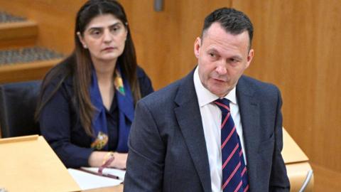 Russel findlay in grey suit and striped tie stands at his podium in the holyrood chamber with female msp seated behind him in black suit and blue scarf