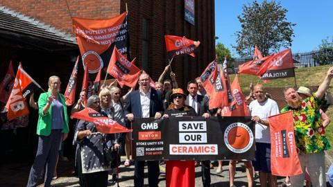 A group of people holding flags and banners with the GMB union logo on them and slogans relating to the ceramics industry. They are standing outdoors, by the entrance to a brick-built building and a grass verge behind them.