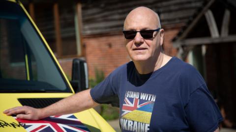Steve Eccleshall stood next to an ambulance with his hand on the vehicle, next to a UK flag. He is wearing a "I stand with Ukraine" tee-shirt which shows both the UK and Ukraine flags.