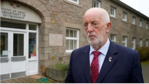 Deputy Steve Luce is wearing a blue blazer and has a red tie on. He is stood in front of the States of Jersey environment department. 