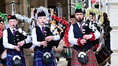 A man and two boys paying the bagpipes, which have been decorated with tinsel, they are wearing kilts - the older man in a red check one, the younger boys in purple, and smart shirts and waistcoasts with purple ties. They also have on some black hats and are marching outside City Hall, a large stone building.
