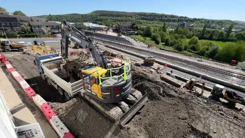 Building works taking place on the railway line near Huddersfield station. There is a digger in the foreground and a railway rack in the background, with moors in the background as well. 