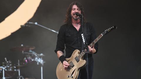 Dave Grohl and Foo Fighters performing on the Pyramid Stage at Glastonbury Festival 2023. He is smiling holding a guitar and has drums in background