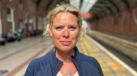 Katie Swinburne looking into the camera with a slight smile. She has blonde hair which is tied back and is wearing a blue jacket, silver necklace and white top underneath. She is standing in a train station on a platform which is out of focus.
