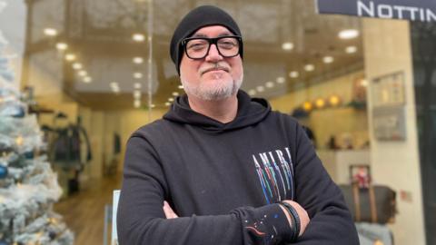 A man wearing a black beanie hat and black hoodie crossing his arms outside a shop window.