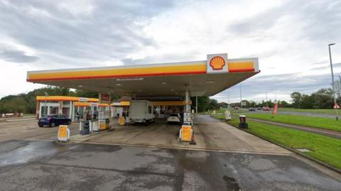 A Shell petrol station next to a dual carriageway. A van and two cars are on the forecourt.