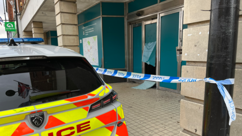 A police car outside a shop that has blue plastic on the windows. One of the windows appears to have been smashed. Police tape has been put in front of the site.