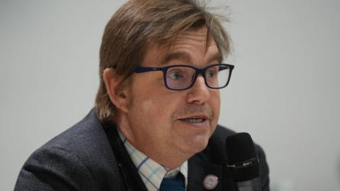 Nik Johnson is looking to the side of the camera while speaking into a microphone. He wears square-framed black glasses and a suit. He has a stubbly moustache and light brown hair.