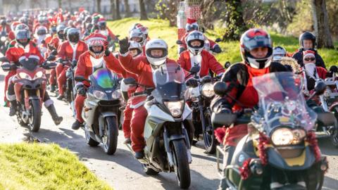 A large convoy of motorcycle riders, with each rider wearing a red and white Santa Claus costume. Some motorcycles are also decorated with Christmas lights and tinsel. 